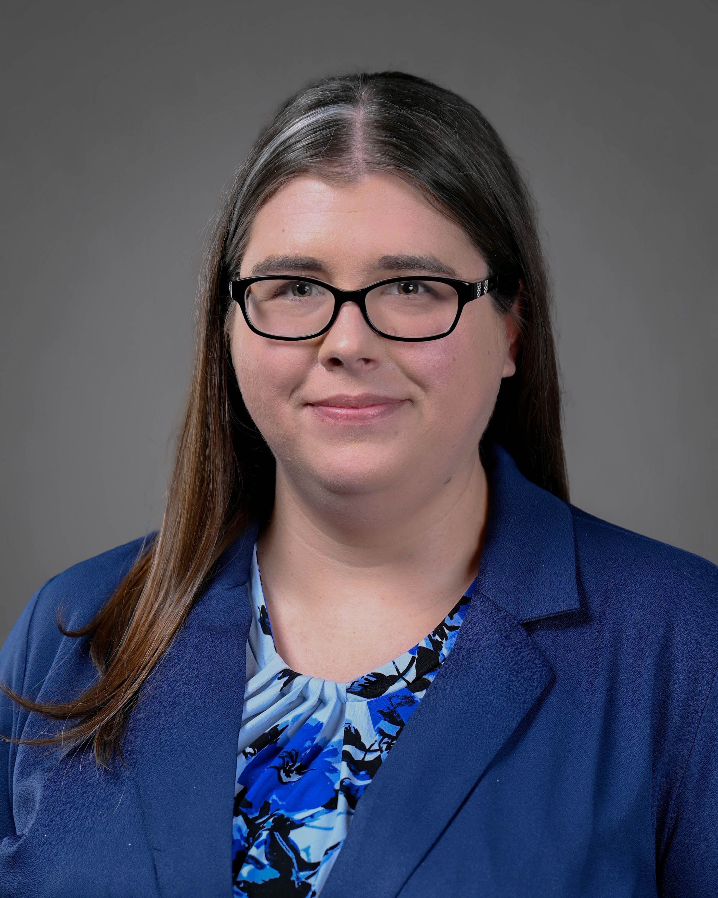 Woman smiling with long dark hair wearing glasses and a blue jacket and blue floral print shirt under.
